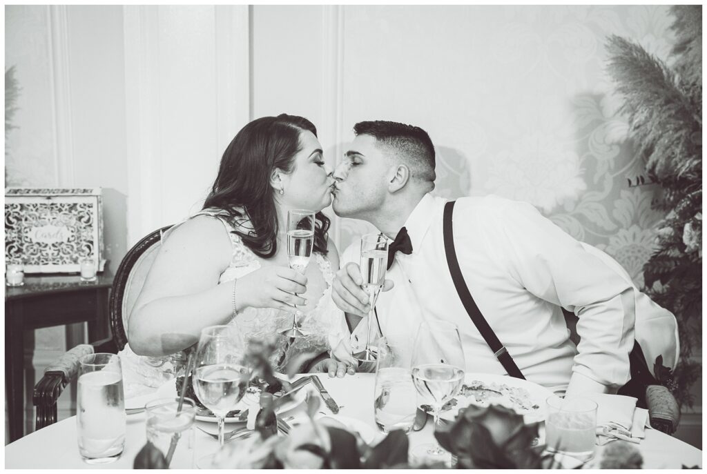 wedding couple sharing a kiss while drinking champagne at the sweetheart table