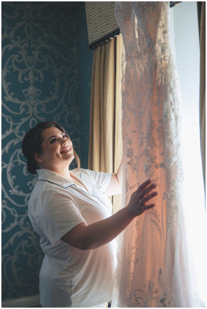 bride standing next to a window with her dress hanging next to her