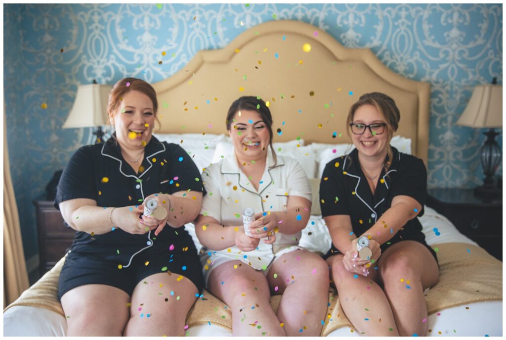 bride and her 2 bridesmaids sitting on a bed wearing matching pajamas and popping confetti