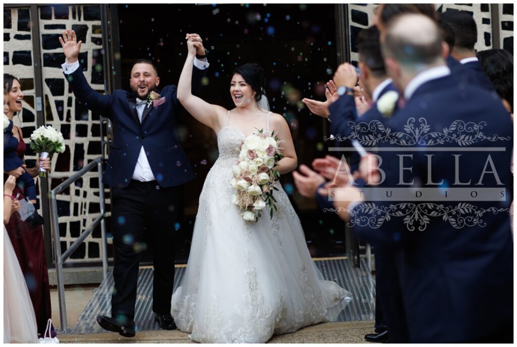 bride and groom cheer as they walk out of the church surrounded by bubbles