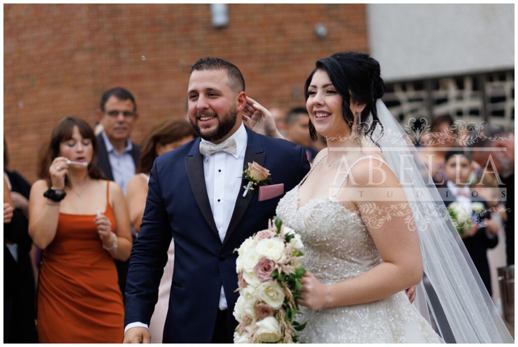 bride and groom cheer as they walk out of the church surrounded by bubbles from guests